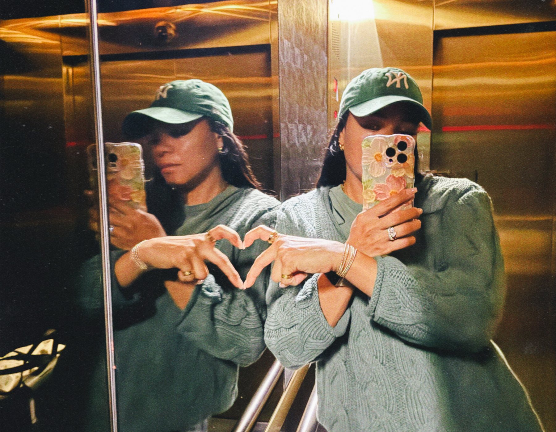 Amanda in an elevator mirror wearing a green baseball cap, making a heart shape with her hands.