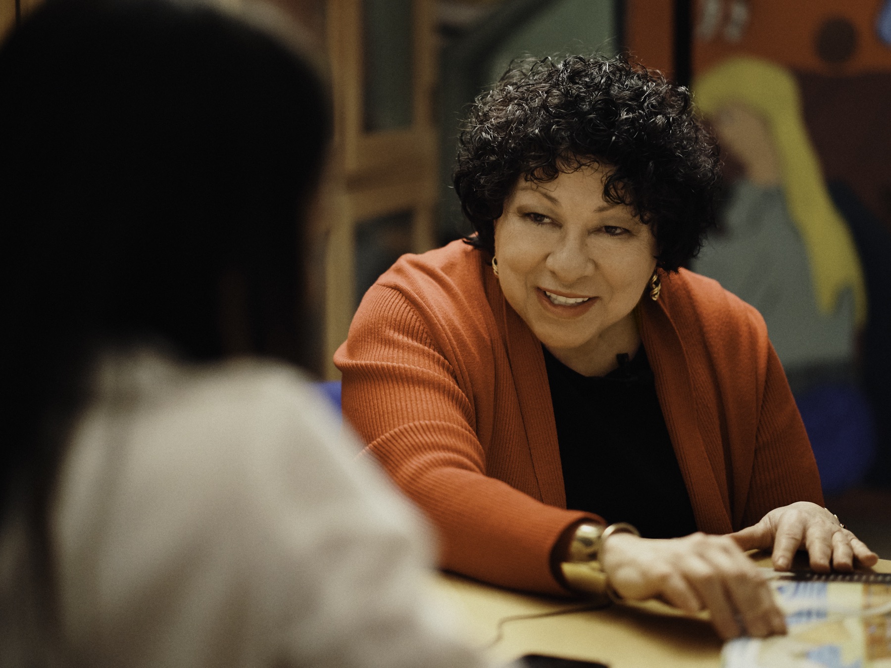Justice Sonia Sotomayor, in a terracotta cardigan, leaning across a table mid-conversation.