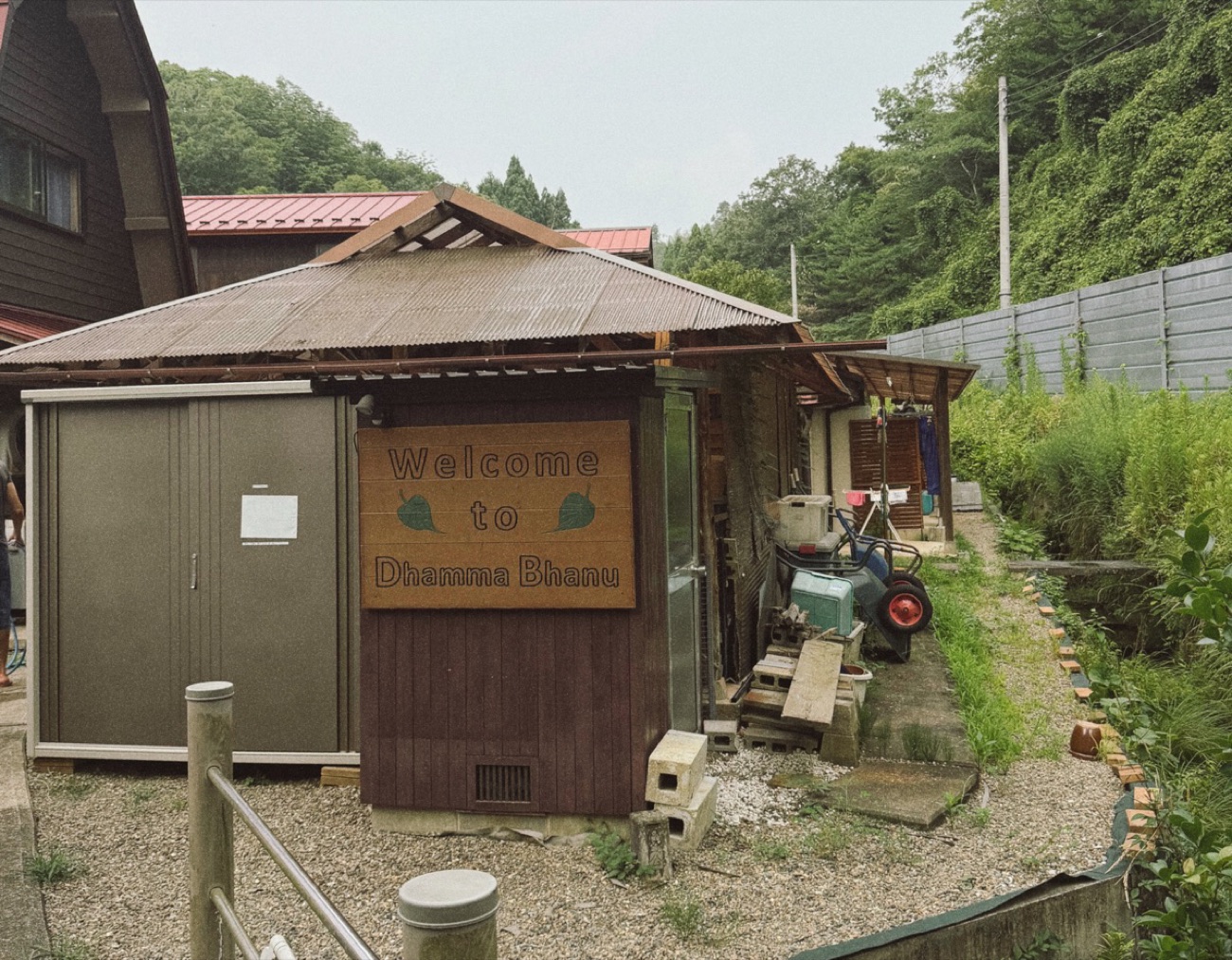 A wooden hand-painted sign reading "Welcome to Dhamma Bhanu" on a small building at the meditation centre, with forested hills behind it.